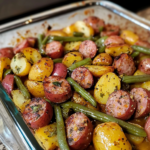 This image shows a close-up of a fork holding a bite of food from a hearty one-pan meal. Speared on the fork are: A small, round baby potato with a golden-yellow hue and a lightly seasoned exterior. A slice of sausage, browned on the edges with a slightly reddish interior. A few green bean segments, cooked until tender but still retaining a vibrant green color. The bite is coated in a light seasoning—likely salt, pepper, and possibly other herbs—evident from the small dark specks visible on the food. Below the fork, on the plate, you can see additional baby potatoes, sausage slices, and green beans arranged in a casual mound. Their surfaces appear lightly glistening, suggesting they have been roasted or baked together, allowing flavors to meld. Sausage, Green Bean & Potato Casserole
